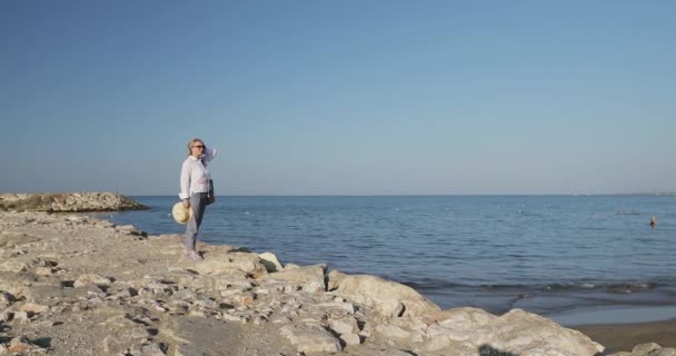 Vieja con estilo en un sombrero se encuentra en una pequeña roca en la playa. — Vídeos de Stock