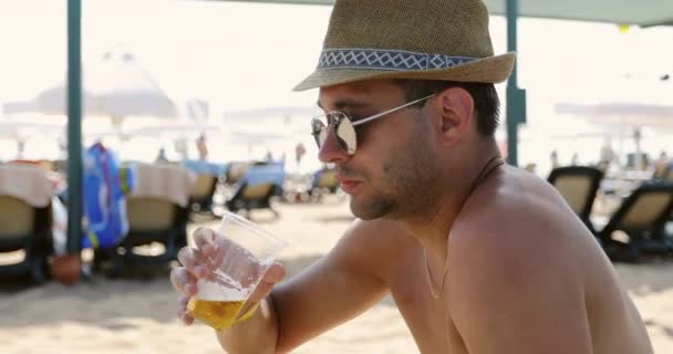 Un hombre con gafas de sol y un sombrero bebe cerveza en la playa. — Vídeos de Stock