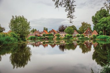 Canal next to rustic houses with bushes and trees in Drimmelen