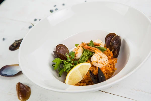 An overhead photo of a seafood risotto on teal textures, with a fork and a spoon, a glass of white wine, and a place for text