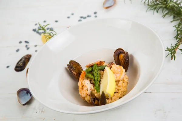 An overhead photo of a seafood risotto on teal textures, with a fork and a spoon, a glass of white wine, and a place for text