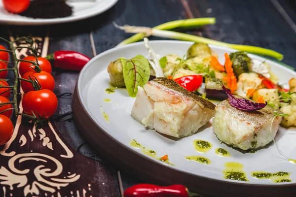 fish with steamed vegetables on a white plate. wooden background decorated with vegetables