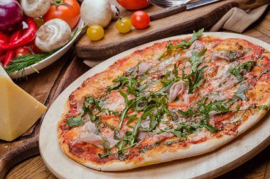 Ahşap bir tahta üzerinde jambon, domates, peynir ve arugula ile İtalyan pizza, arka plan sebze ve çatal ile dekore ahşap