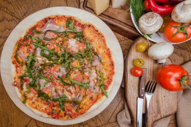 Ahşap bir tahta üzerinde jambon, domates, peynir ve arugula ile İtalyan pizza, arka plan sebze ve çatal ile dekore ahşap