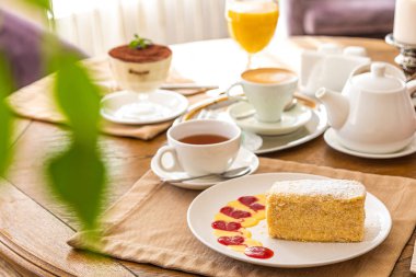 Servis masasında beyaz bir tabakta bir dilim Napolyon keki. Arka planda çaydanlık, çay, meyve suyu var..