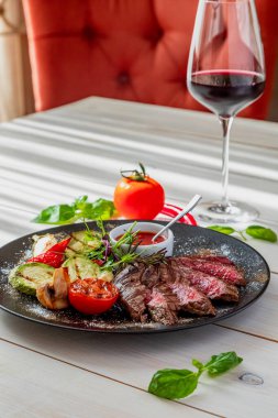 Orta pişmiş biftek ve ızgara sebze. Tabak domates sosu ve bir bardak kırmızı şarapla servis edilir. Tabak beyaz bir tabakta, doğal ışıkta.