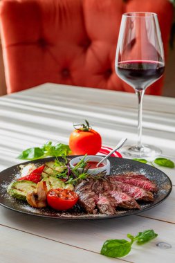 Orta pişmiş biftek ve ızgara sebze. Tabak domates sosu ve bir bardak kırmızı şarapla servis edilir. Tabak beyaz bir tabakta, doğal ışıkta.