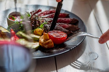 Kadın eli biftek kesiyor. Orta pişmiş biftek ve ızgara sebze. Tabak domates sosu ve bir bardak kırmızı şarapla servis edilir. Tabak beyaz bir tabakta, doğal ışıkta.