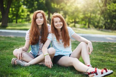 Kızıl güç. İki güzel kardeş parkta çim üzerinde oturan ve sevinçle gülümseyen kamera, zevk sıcak güneşli bir gün, kız kardeş, yakın ilişki yaşamak yaslanmış, Kızıl saçlı