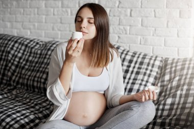 Evde istirahat ve rahatlatıcı vücut kremi kokulu kokusu zevk, kanepe oturma odası, kapanış gözleri ve koklama hoş kokulu, bebek doğmak bekleyen oturan genç hamile kadın