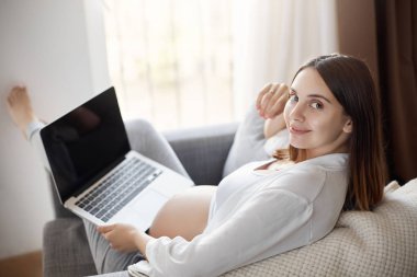 Soğutma ve evde rahatlatıcı, süre annelik hakkında video izlerken, koltukta yalan ve laptop kucağa, elinde koca bir bardak süt soran hamile kadın kamera hoş ifade ile gülümseyen