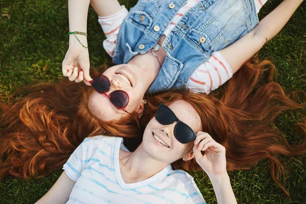 Sinto-me como chefe. Retrato de encantador e despreocupado ruiva irmãos femininos com sardas, deitado na grama no parque e vestindo óculos de sol na moda enquanto rindo e sorrindo, discutindo a forma de nuvens — Fotografia de Stock