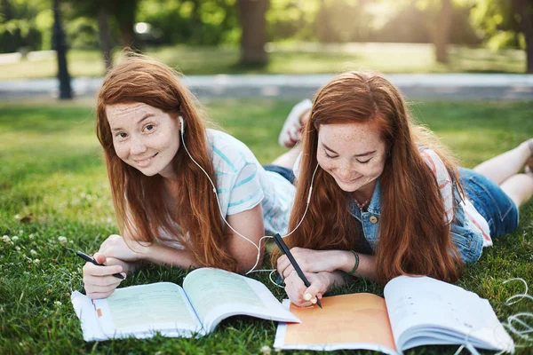 Pekerjaan rumah yang kau bercanda. Dua gadis cantik dengan rambut merah berbaring dan menggigil di rumput selama waktu luang, melakukan pekerjaan rumah, kakak membantu saudara kandung dengan pelajaran, tersenyum dan tertawa — Stok Foto