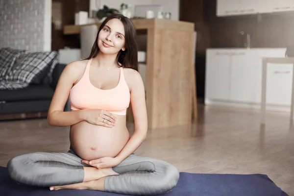 Lodret skud af flot sund og aktiv gravid kvinde i sports bh og leggings, holder håndfladerne sammen og sidder i yoga på rullemåtte derhjemme, mediterer, bliver klar til barsel Stock-foto ©