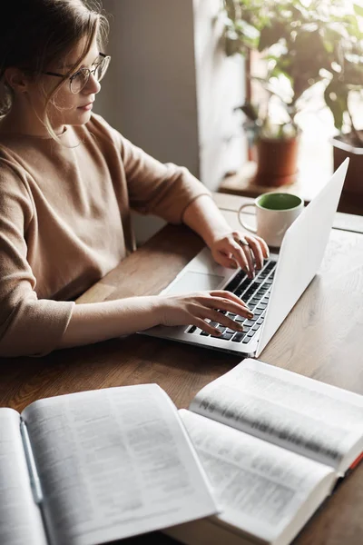 Tiro vertical de mujer atractiva con estilo determinado y centrado en gafas, sentado en la mesa de madera, trabajando con el ordenador portátil y libros, escribiendo carta al cliente, buscando información para el futuro proyecto — Foto de Stock