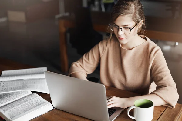 No hay tiempo para distraerse, hay que trabajar duro. Encantadora empresaria ocupada con gafas y ropa elegante, sentada cerca de diccionarios y computadora portátil, trabajando con Internet, bebiendo té caliente para concentrarse —  Fotos de Stock