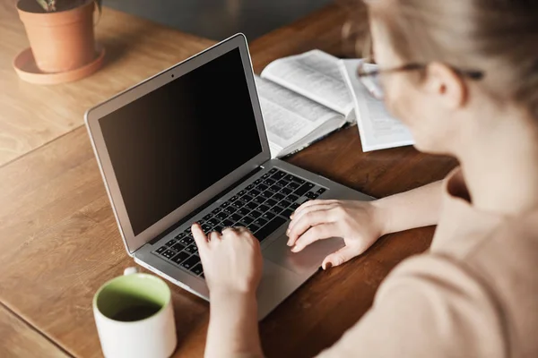 Primer plano de ocupada mujer de negocios guapa y enfocada, sentada en la cafetería, trabajando con la computadora portátil, buscando información, haciendo investigación para el proyecto universitario, bebiendo té, revisando información en libros — Foto de Stock