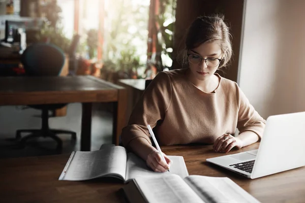 You cannot distract girl with anything. Portrait of charming focused caucasian female student in glasses, writing with pen in notebook, working with laptop, gathering information from internet — Stock Photo, Image