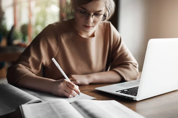 Close-up shot of hardworking attractive female entrepreneur in glasses, writing in notebook, checking spelling in dictionary, making homework for university, using laptop to browse in network — Stock Photo, Image