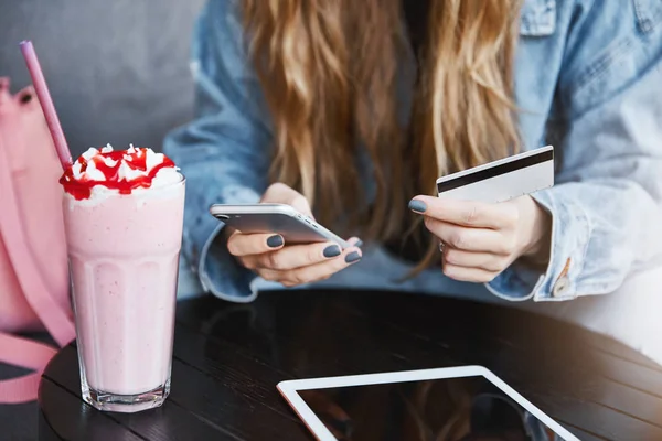 Schnappschuss einer jungen Frau mit blonden Haaren, die Smartphone und Kreditkarte in der Hand hält, Bankkontonummer eingibt, um den Kontostand zu überprüfen, Cocktail trinkt und Nachrichten in digitalem Tablet liest — Stockfoto