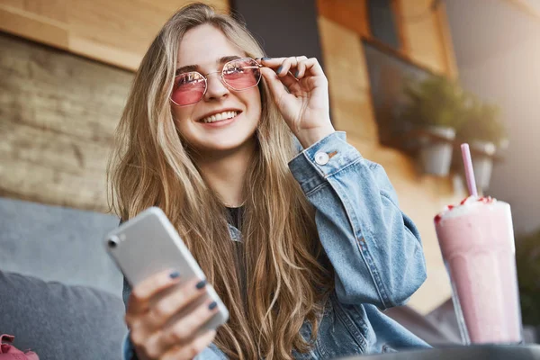 Atraktivní a radostné kavkazské blondýnka v módní sluneční brýle, dotýkat okraj brýlí a široce se usmívala a zíral na městských ulic, držení smartphone, chatování během oběda, pít koktejl — Stock fotografie