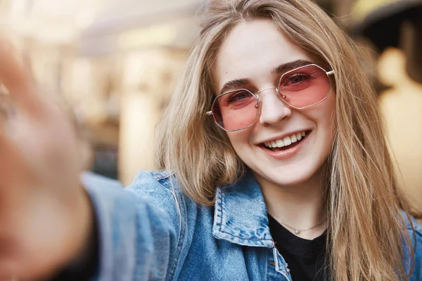 Selfie je čas. Detail z okouzlující sebejistý a stylové kavkazské blondýnka v trendy růžové brýle a denim, vytáhl ruku směrem k fotoaparátu pořizovat Foto, usmíval se radostně, aktualizace profilu zastřelil — Stock fotografie