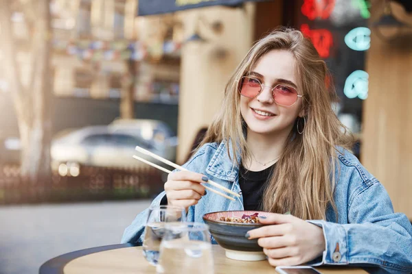 Waktu untuk meningkatkan energi, kreatif dan wanita bisnis muda berhenti membeli selama makan siang di restoran Asia, memegang mangkuk dengan ramen dan sumpit, menatap dengan senyum lebar di kamera, makan makanan pedas — Stok Foto