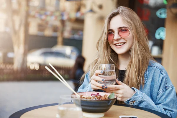 Dobře vypadající šťastné mladé městské studentka v džínové sako přes černé tričko, držení sklenice vody a hledí s širokým úsměvem stranou, jíst v asijské restauraci s hůlkami — Stock fotografie