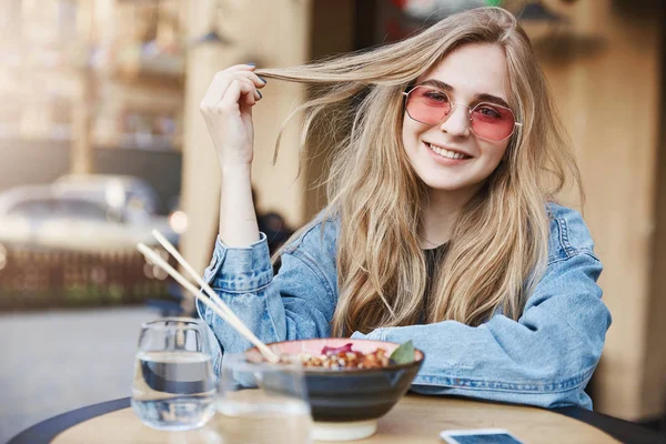 Foto luar ruangan dari wanita atraktif dengan rambut yang adil, bermimpi untuk menjadi blogger fashion, berdiri di restoran jalanan Asia, makan soba dengan sumpit, tersenyum dan bermain dengan helai rambut — Stok Foto