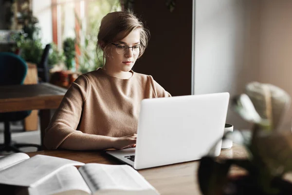 Werk-, lifestyle- en business concept. Knappe gerichte Europese vrouw in trendy bril zitten in Cafe in de buurt van laptop, werkt op laptop, omringd met boeken, het maken van notities voor les met kinderen — Stockfoto