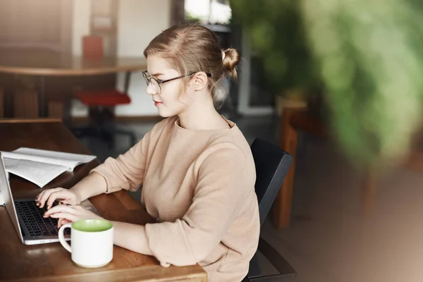 Perfil tiro de atraente focado e determinado professor freelance feminino em óculos e roupas da moda, trabalhando em laptop, olhando para a tela enquanto prepara o material de trabalho, bebendo chá no café — Fotografia de Stock