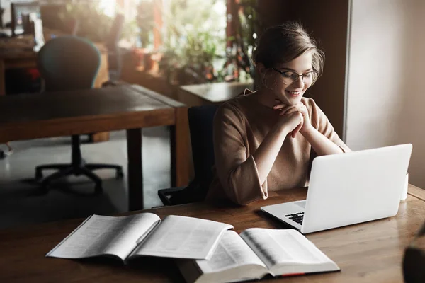 Mooie vrouw in glazen praten met een vriend via laptop, breed glimlachend op scherm, hand in hand onder de kin, zitten in de ruimte van de kunst, pauze studeren, omsingeld met boeken en notities — Stockfoto