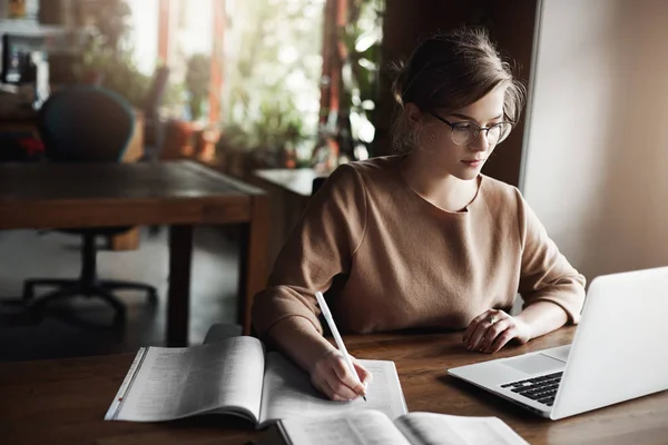 Linda mujer trabajadora haciendo notas, copiando información del sitio de Internet, utilizando el ordenador portátil mientras trabaja en el ensayo de la universidad, sentado en la cafetería, estudiando y tomando café, usando gafas de moda — Foto de Stock