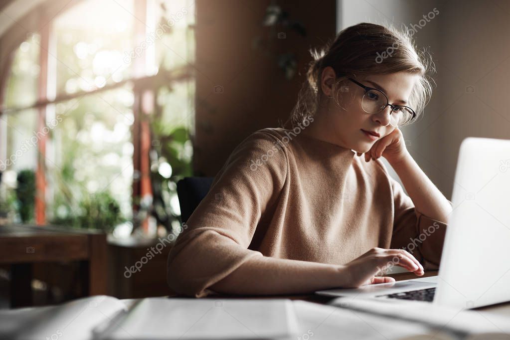 Good-looking young female freelancer tutor sitting in cozy cafe, leaning on hand while checking schedule via laptop, gazing at screen with focused expression, working, preparing material, making notes