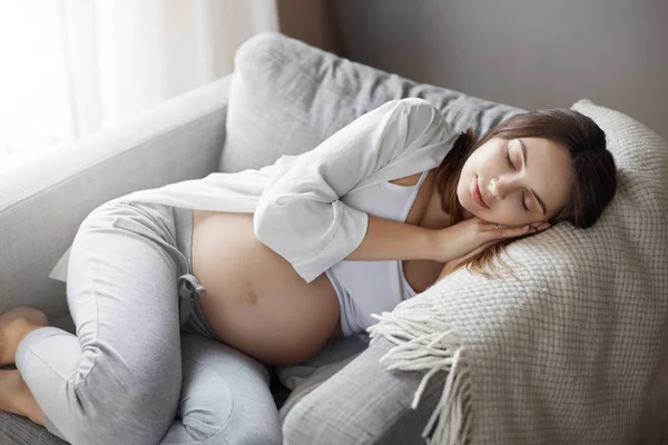 Durmiendo y soñando con el bebé. Retrato de encantadora hembra embarazada tierna acostada en el sillón, tomando siesta y sonriendo, teniendo un sueño agradable y lindo, sosteniendo las palmas bajo la cara — Foto de Stock