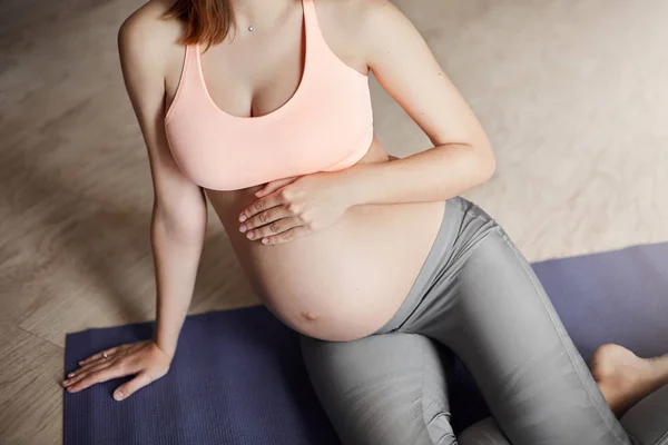 Cropped shot of tender caring pregnant female in leggings and pink sports bra, touching belly while thinking about future baby be born, lying on roll pad at home, taking rest after breathing exercises