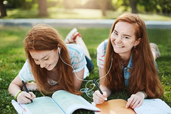 Припиніть обманювати і робіть домашнє завдання. Дві розслаблених і безтурботних рудих сестер з веснянками, лежачи на траві в парку, пишучи нотатки для університету, ділячись навушниками, слухаючи пісні разом, сміючись — стокове фото