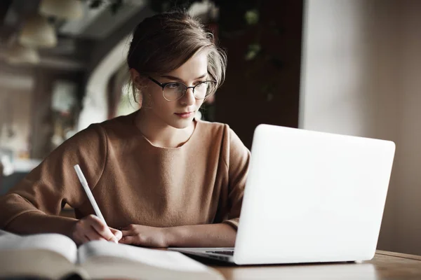 Mulher europeia de boa aparência criativa com cabelo claro em óculos da moda, fazendo anotações enquanto olha para a tela do laptop, trabalhando ou se preparando para reunião de negócios, sendo focada e trabalhadora — Fotografia de Stock