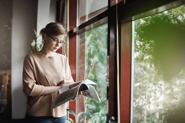 Woman likes spend time with reading. Indoor shot of cute creative and smart female in trendy outfit and glasses, holding magazine and turning pages while standing near window, taking bread from work