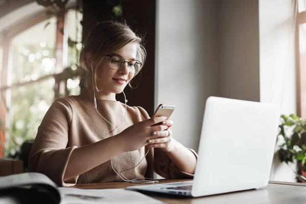 Woman studying and preparing project via laptop, wearing earphones, listening music joyfully, holding smartphone and reading answear on message, being intrigued with future date, taking break — Stock Photo, Image