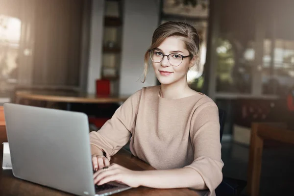 Creative and attractive female graphic designer working under project while sitting in cafe with laptop, gazing at camera with slight tired smile, arranging meeting via notebook with clients — Stock Photo, Image