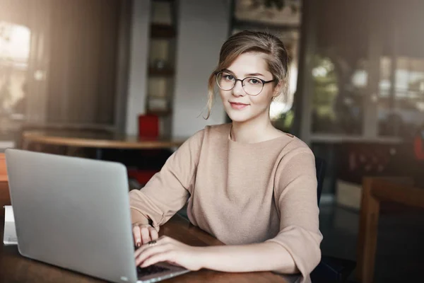 Desainer grafis perempuan kreatif dan menarik bekerja dalam proyek sambil duduk di kafe dengan laptop, menatap kamera dengan senyum yang sedikit lelah, mengatur pertemuan melalui notebook dengan klien Stok Gambar Bebas Royalti