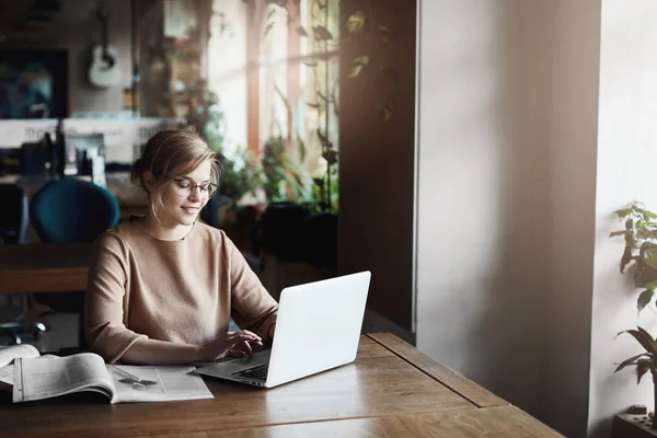 Businesswoman finally found time to answer on customer questions, sitting in stylish office, typing message on laptop, working with books and dictionaries while preparing project for company director Stock Photo