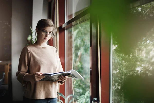 Indoor shot of good-looking serius rekan kerja wanita european di trendi dan nyaman pullover, memegang majalah dan menatap dengan mimpi ekspresi kamera, berdiri di dekat jendela, bermimpi dan santai — Stok Foto