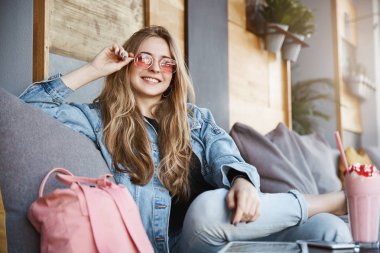 Çekici ve neşeli sarışın kadın iş arkadaşınız kaygısız poz Divan Cafe, arkadaşları ile soğutma sırasında oturan kot ceketli RIM güneş gözlüğü dokunmaktan ve geniş sevinçle gülümseyen