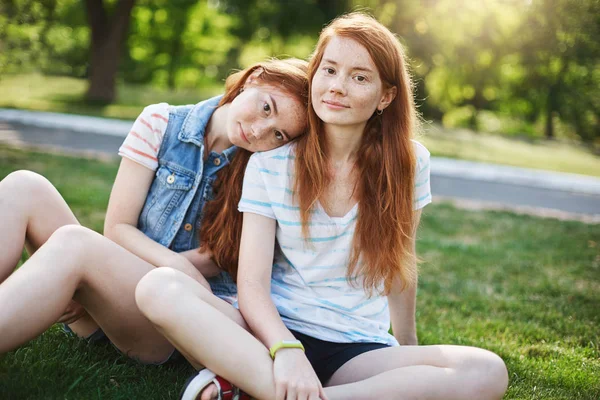 Hon täcker alltid min rygg. Två charmiga rödhårig kvinnor i casual kläder sitter på gräset i parken, Syster lutar på flicka axel och ler mot kameran, att vara avslappnad och glad att ha varandra — Stockfoto