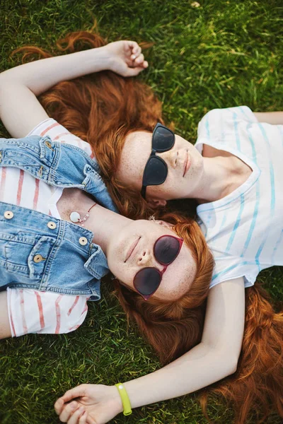 Vertikala skott av vackra avslappnad och glad flickor med naturlig ingefära hår och söta fräknar liggande på gräset, vidrör varandra huvuden, bära coola solglasögon, kylning under solen i varm sommardag — Stockfoto