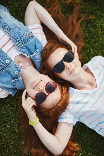 Os gémeos são os melhores. Tiro vertical de duas belas irmãs com cabelo de gengibre e sardas, deitado na grama e sorrindo com expressão relaxada, tocando rostos, expressando cuidado um com o outro — Fotografia de Stock