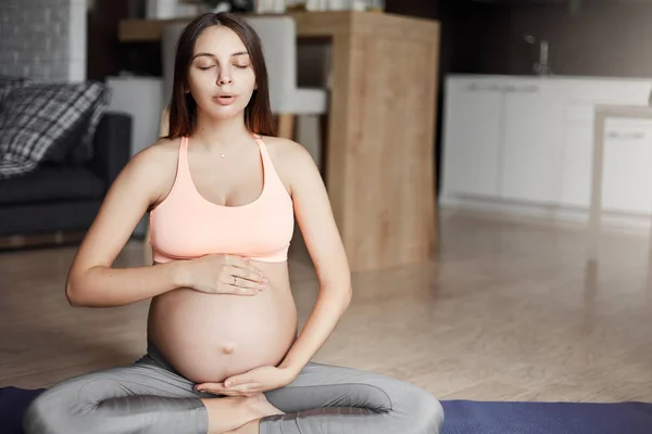 Mulher grávida atraente fazendo exercícios respiratórios para levar um estilo de vida ativo e saudável durante a gravidez, sentado na almofada de rolo em pose de ioga, tocar na barriga, lábios dobráveis e fechar os olhos — Fotografia de Stock