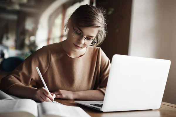 O prazo aproxima-se, mas a rapariga nunca perde a paciência. Mulher focada e trabalhadora em óculos da moda concentrando-se em escrever ensaio, sentado em um café acolhedor perto do laptop, trabalhando e fazendo anotações com cuidado — Fotografia de Stock